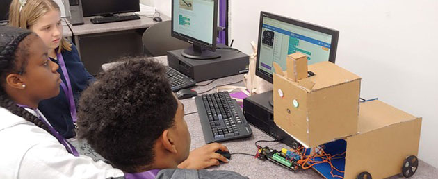 multi ethnic middle school children sitting at computer with a robot made of cardboard to the right of the computer.