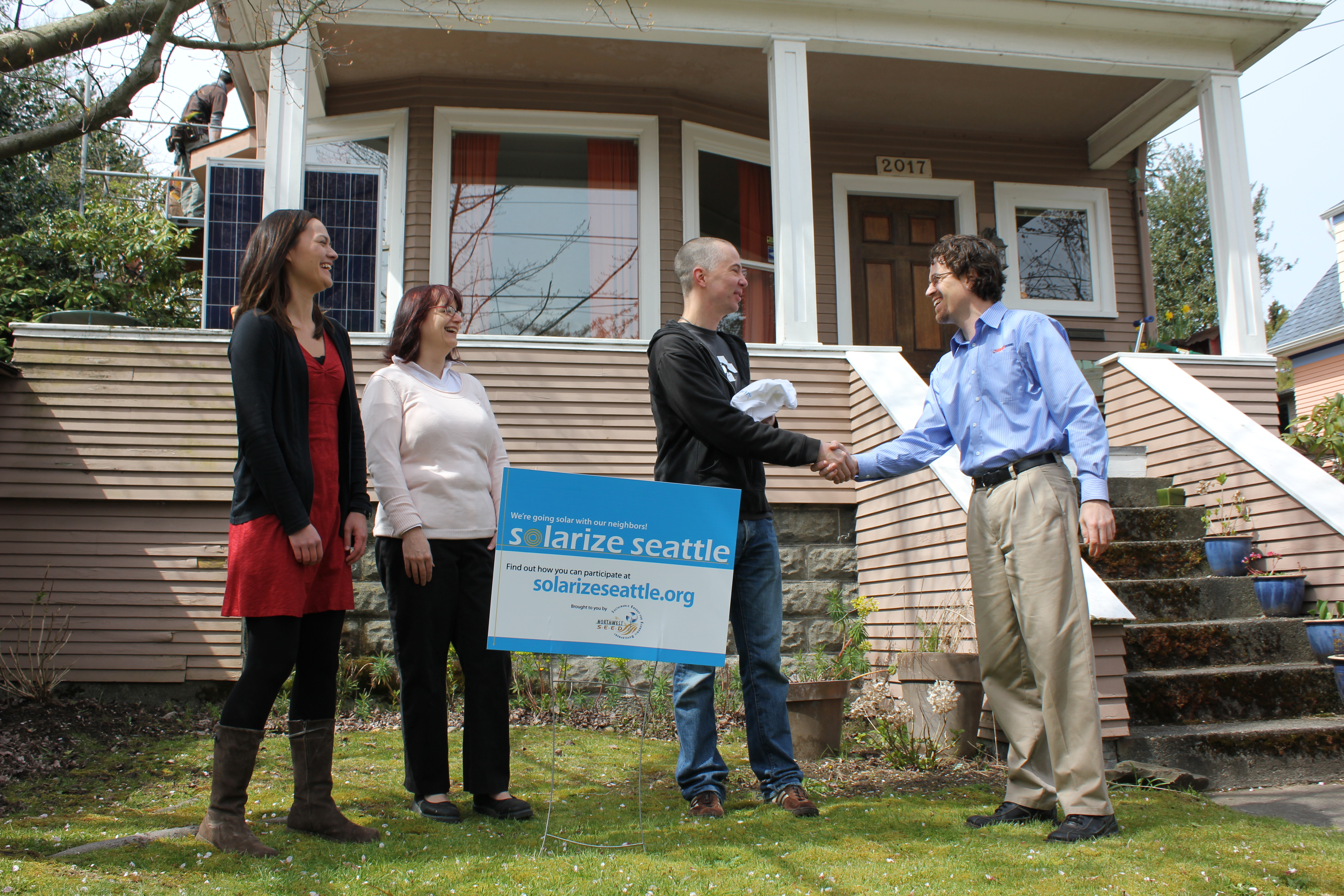 two male and two female adulst in fromnt of beige colored house. The men are shaking hands in from of a Solarize Seattle lawn sign. All people are smiling.