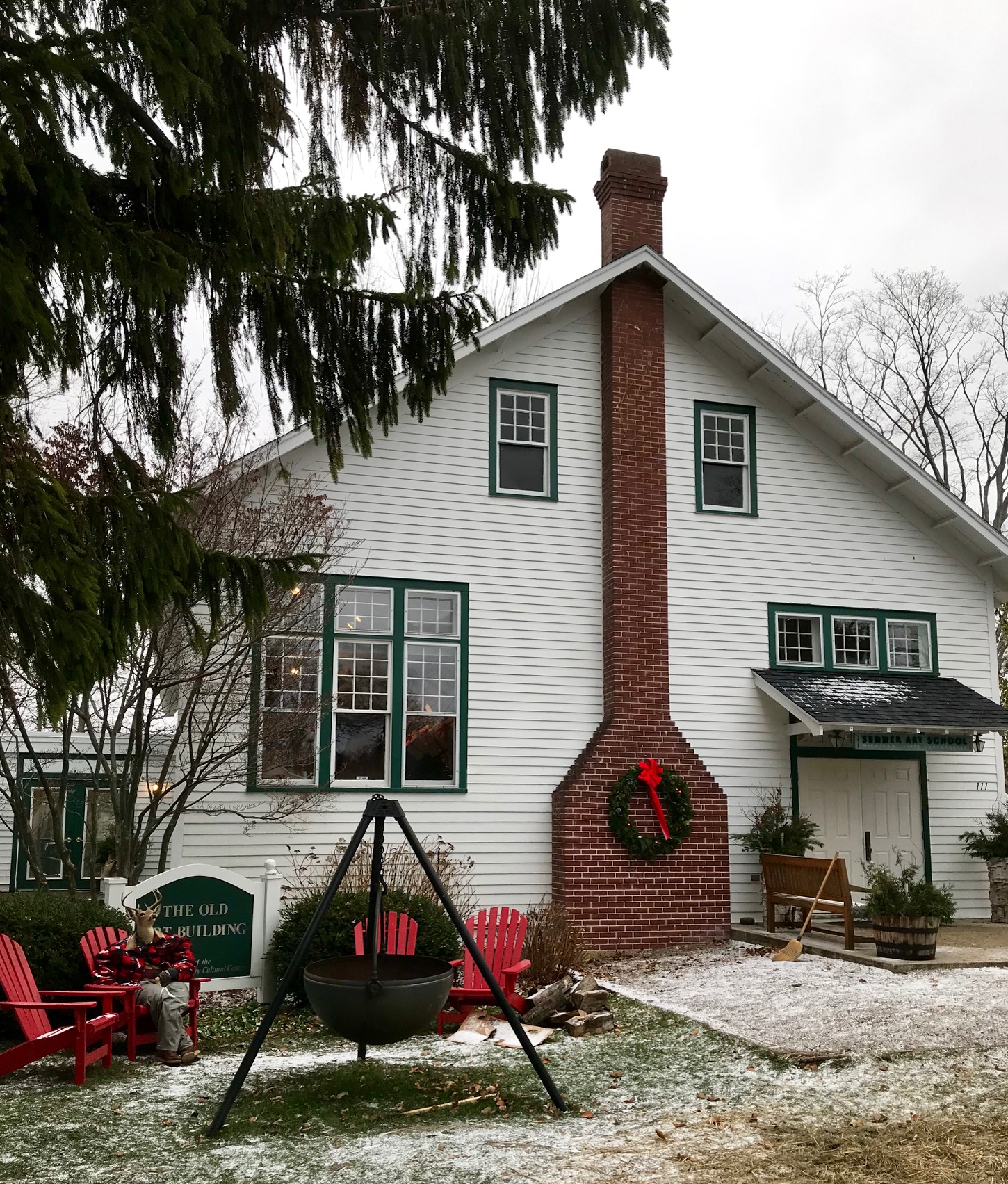 side view of a colonial building/house. Brick chiminey in the milddle, with Christmast wreath. Red adirondak chairs around cauldron fire pit.