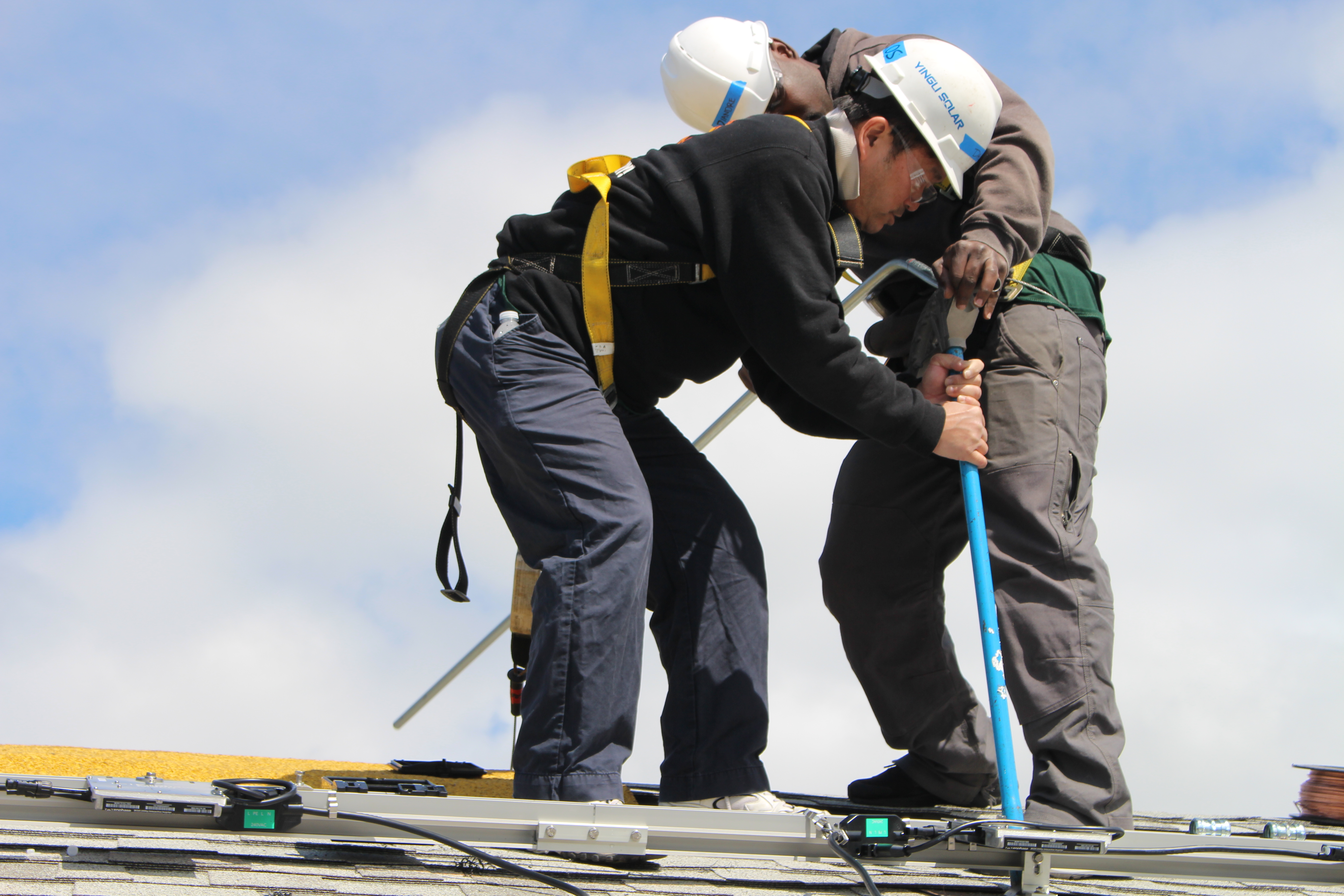Two men in hard hats standing on a roof, installling solar panel hardward.