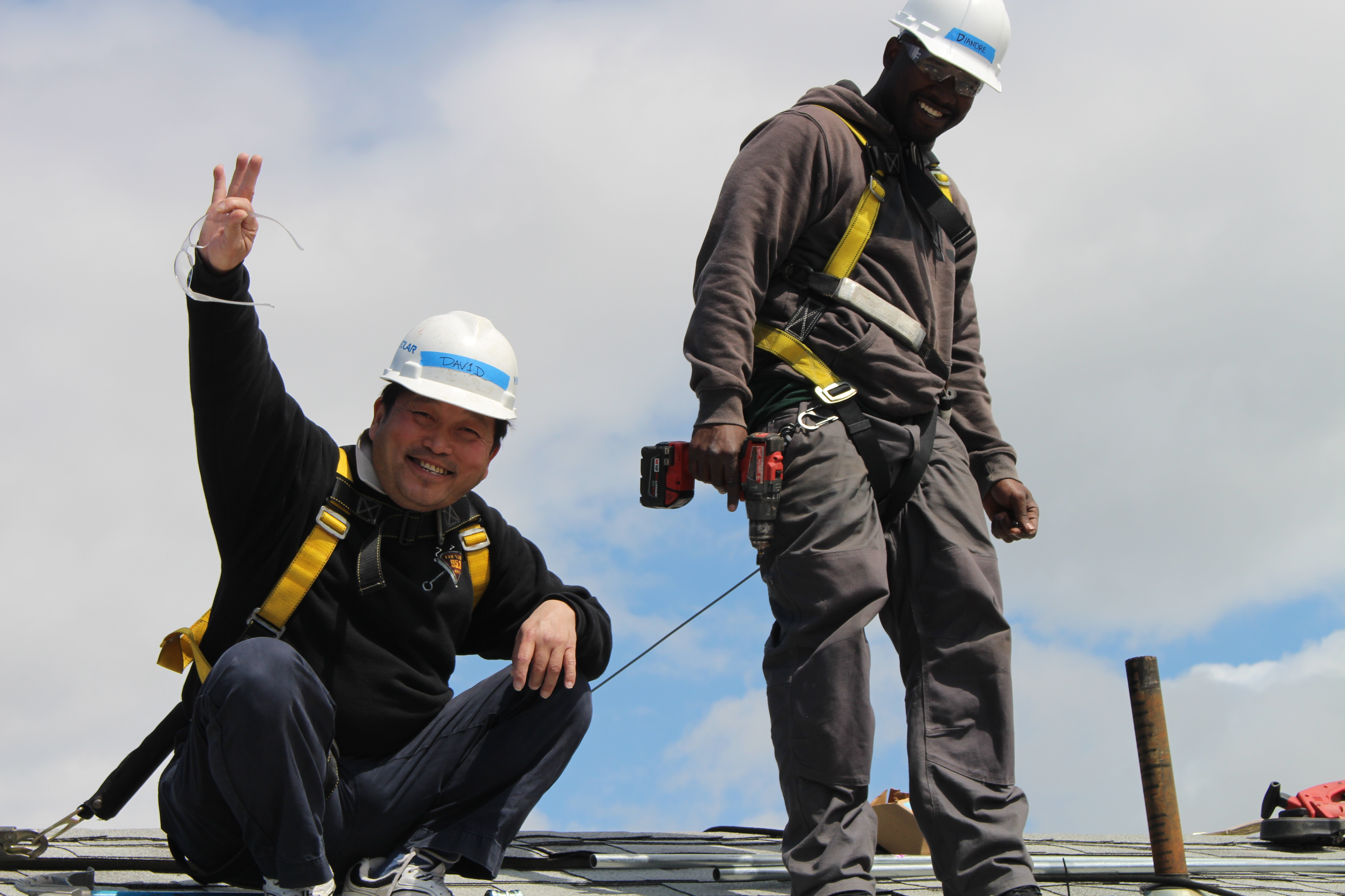 One hispanic and one african american males, on a roof wearing hardhats. One man is waving and the other man is holding a drill. Both are wearing saftey harnesses and smiling.