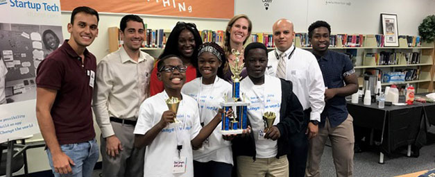 three african american middle school children, two boys, one girl with six adults gathered for a photo, children are holding trophies