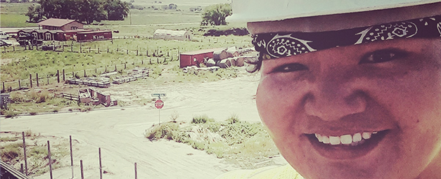 American Indian woman in hard hat and bandana, on roof, taking a selfie, with farmland, farm buildings and dirt roads in the background.