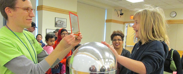 Man wearing glasses and holding a mirror whild a woman puts her hand on a plasma ball, to see the effects of static electricity in the mirror. Other adults and children in the backgound smile and look on.