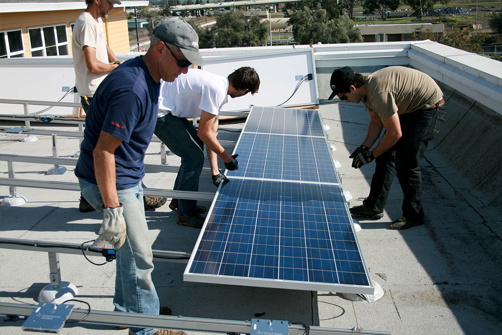 four men on the roof of a commercial building. All are wearing work gloves and working on the installation of solar panes.