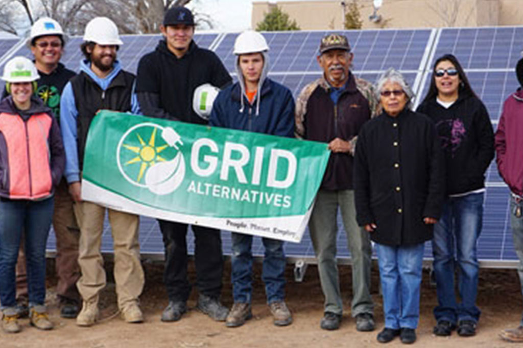 nine adults of varying ages standing in front of a solar panel installation. Two men are holding a green banner with the Grild Alternatives logo.