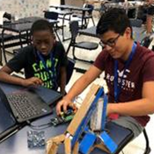 Two middle school boys working at a table on a cardboard robot with laptop on the table.