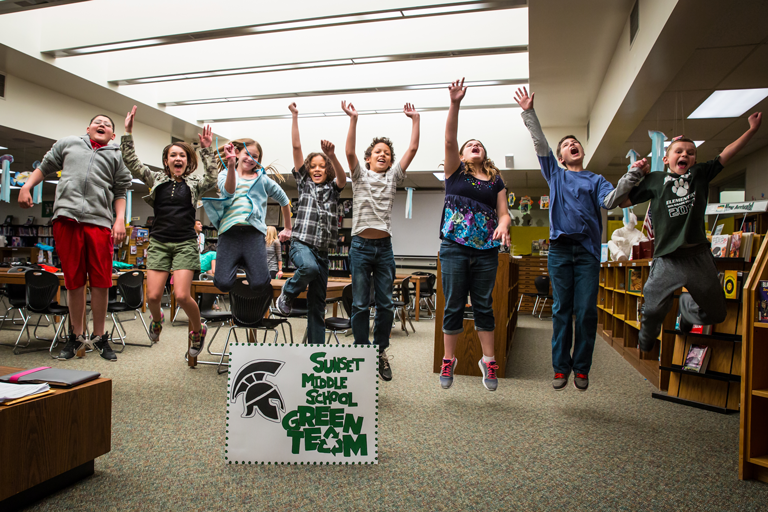 eight middle school childen in a library jumping for joy with arms up in the air. Sign with 
