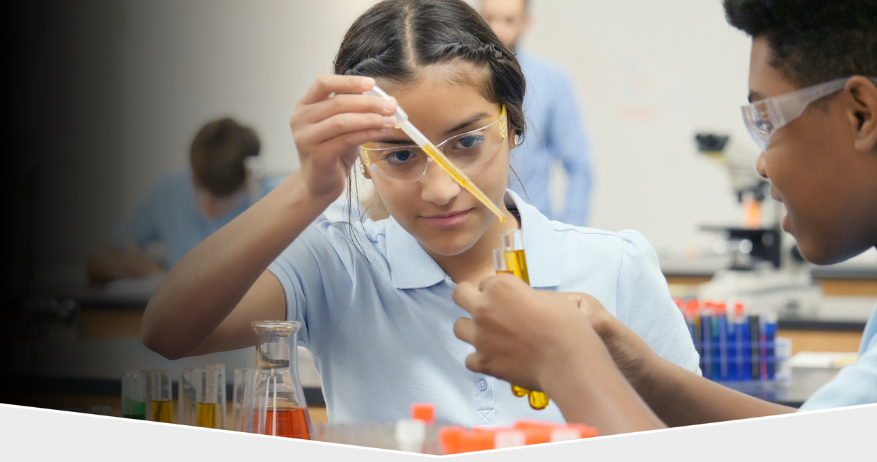 Ethnically diverse middle school boy and girl, wearing safety glasses, working in a chemisty lab. girl is using an eye-dropper to add yellow liquid to a beaker.