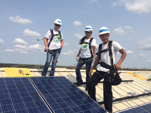 Three women in hard hats and Grid Alternatives shirts, saftey harness, carrying tools, working on a roof installing solar panels.