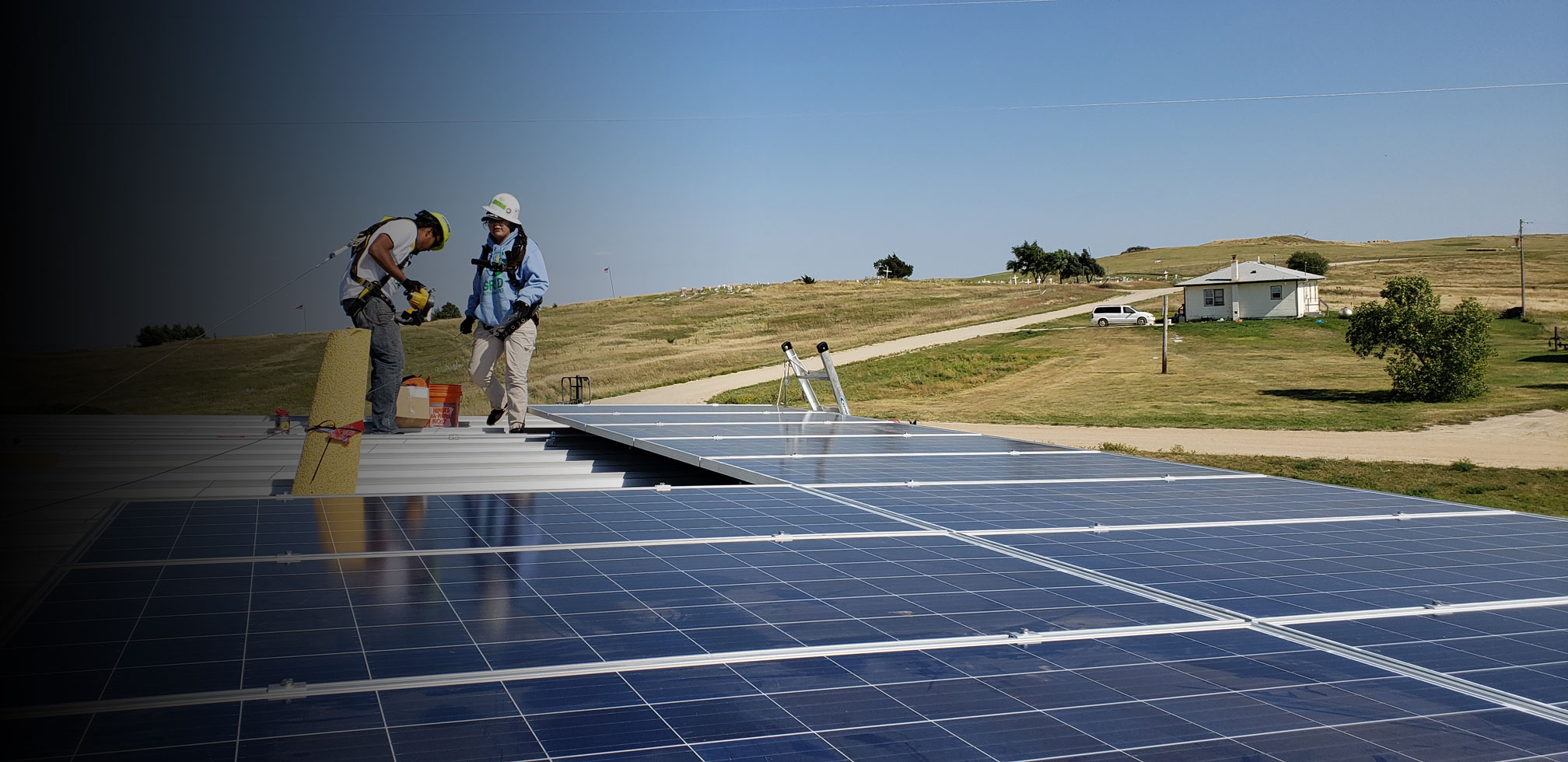 two men in hard hats and safety harnesses, working on a flat roofed building in the middle of rolling hills, installing solar panels. dirt road, a few trees and blue sky in the background.