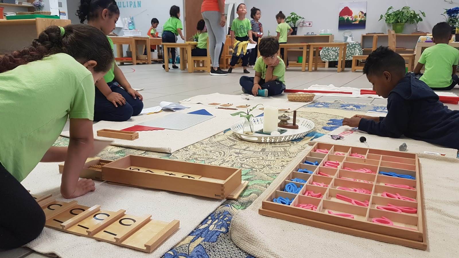 ten young elementary school children, ethincnally diverse, working on projects in their classroo,m. Children in foreground are sitting or laying on the floor working with woodent blocks, shapes and numbers