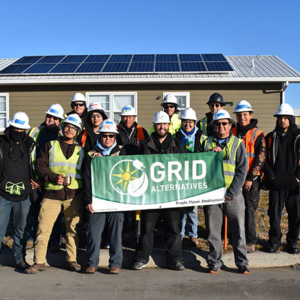 Diverse group of male and female workers in hard hats and protective gear holding a green Grid Alternatives sign in front of a brown house with solar panels on the roof