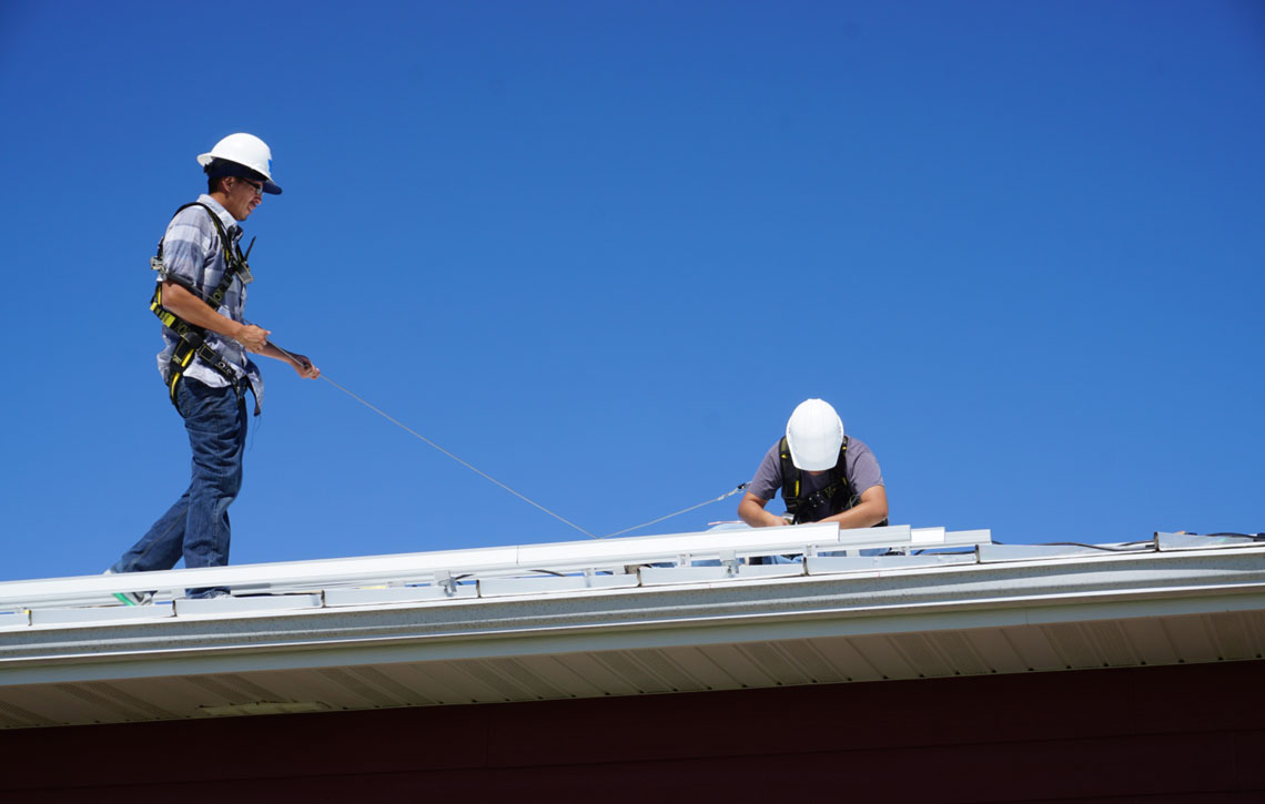 Two men in hard hards and protective equipment on a roof installing solar panels
